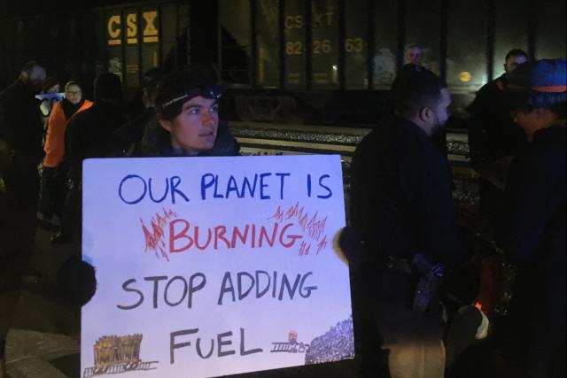 Activists stand beside a coal train at night; one holds a sign reading "Our planet is burning—stop adding fuel"