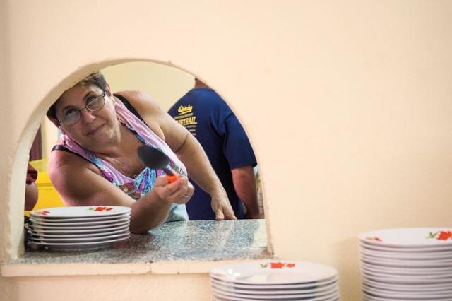 A smiling Cuban woman holding a serving spoon peers through a kitchen pass-through