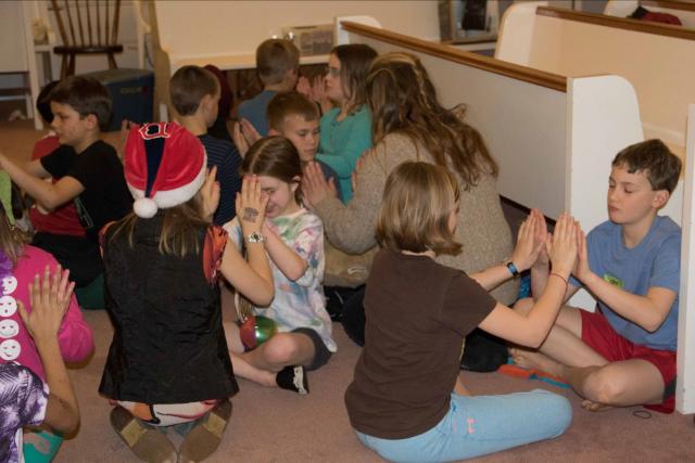 Several children, sitting in pairs facing one another with palms nearly together