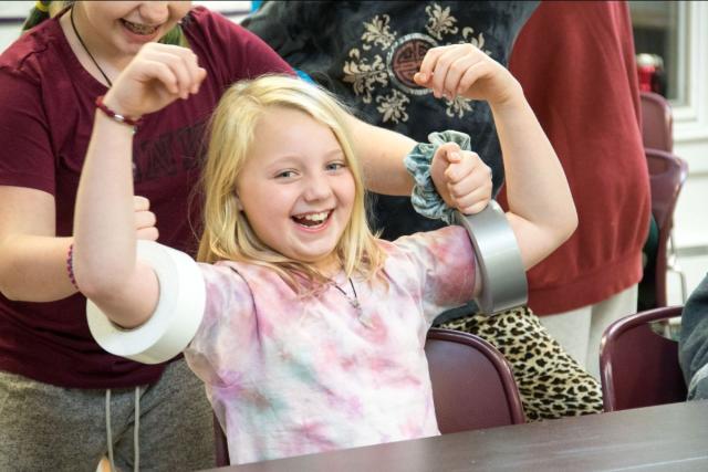 A young girl with blond hair flexes her arms, which have rolls of duct tape on them