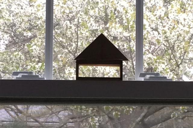 A view of a window which has a small wooden model of a building perched on the sill