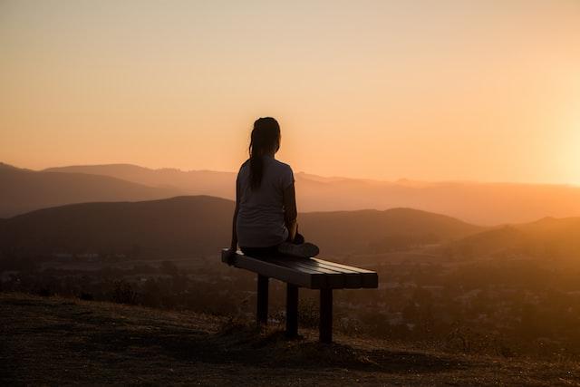 Image of a woman sitting on a bench with a sweeping landscape before her, all bathed in low rays of the sun.