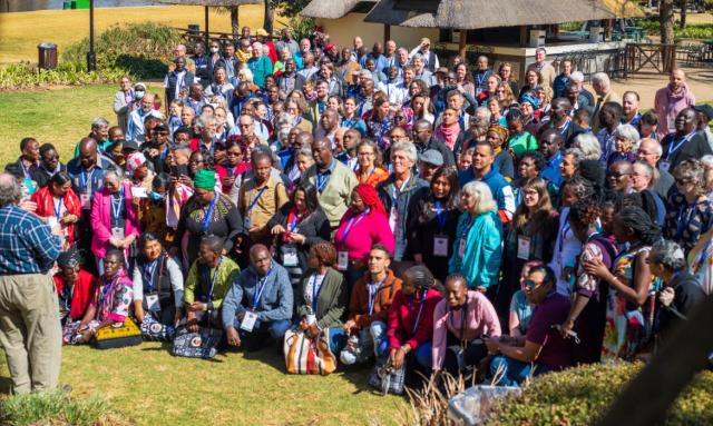 The image shows a large group of people of different races and ages, outdoors with a grass-roofed structure in the background