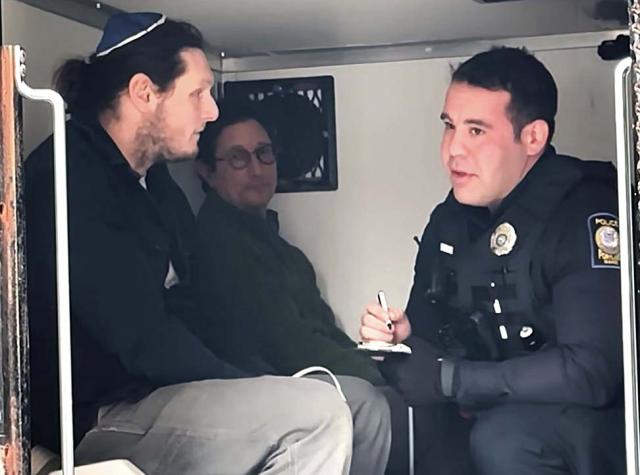 Two men sit in the back of a police van with a male officer