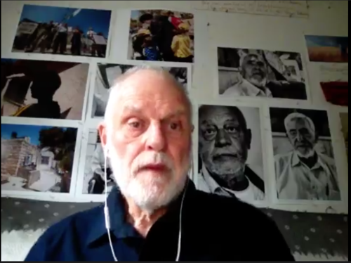 Image of a man with white hair and beard, with photographs on the wall behind him.