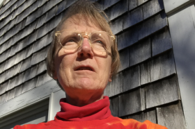A photo of a woman with short, gray hair, wearing glasses and a bright orange sweatshirt