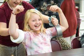 A young girl with blond hair flexes her arms, which have rolls of duct tape on them