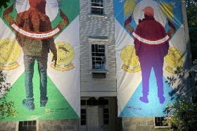 Two large banners with artwork by immigrants to the U.S. hang on the side of a meetinghouse