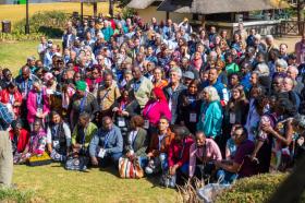 The image shows a large group of people of different races and ages, outdoors with a grass-roofed structure in the background