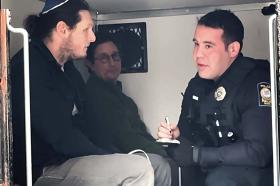 Two men sit in the back of a police van with a male officer
